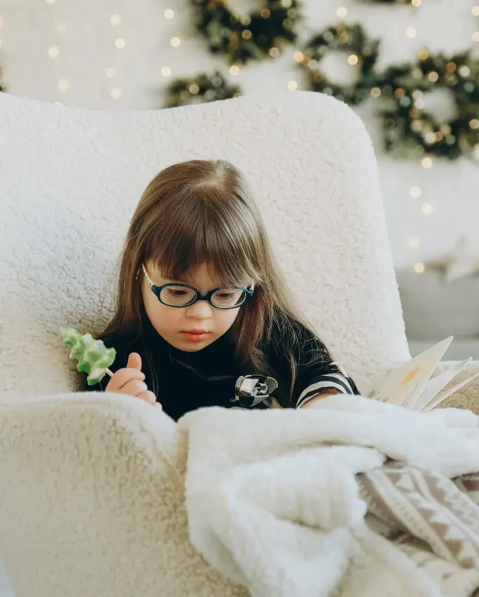 Young girl with Down's Syndrome reading a book in a cozy blanket and chair in front of Christmas decorations.