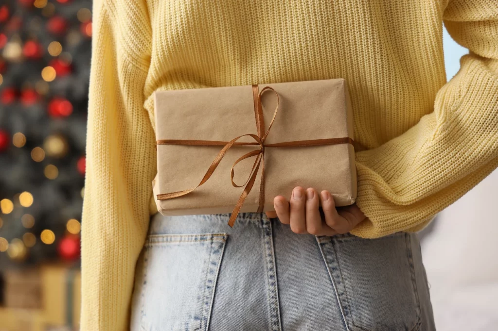 Woman in a yellow sweater holding a brown paper wrapped gift behind her back